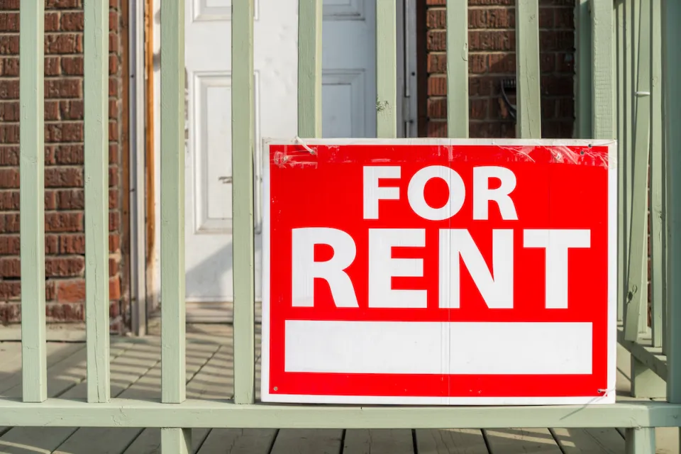 Close-up of red For Rent sign with white text on porck of brick building.